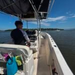 Person standing at the boat’s helm, steering controls visible, with towels on the seat and gear nearby against blue water and distant sailboat on a sunny day.