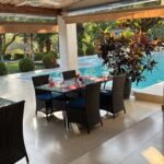 Outdoor poolside dining area with glass table, wicker chairs, red place settings, and a ceiling fan under a thatched roof.”,