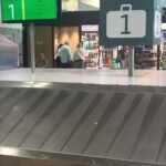 Baggage carousel in an airport with a large '1' sign and green flight information screens in the background, with several passengers nearby.