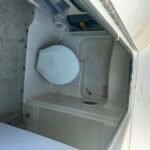 Open top-loading washing machine with visible drain hose and yellow connector, sitting on a dirty floor.