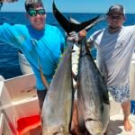 Two men on a boat proudly hold up three large fish they caught, with blue ocean behind them.