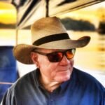 Older man wearing a straw hat and dark shirt, sunglasses, outdoors on a sunny boat or lakeside setting