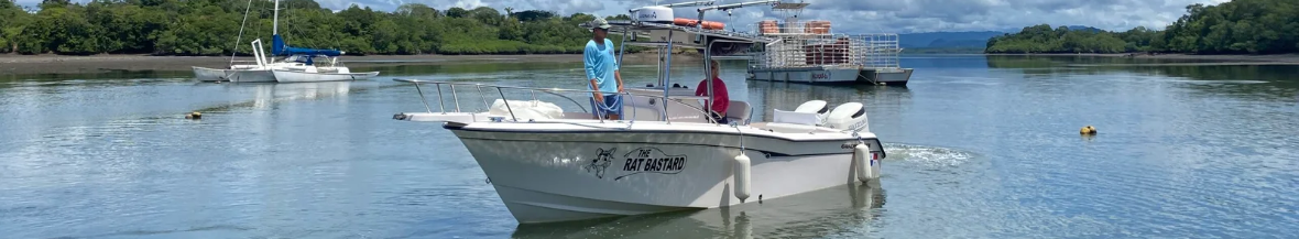 rbbb - Panama Sport Fishing Charters Powerboat named The Rat Bastard gliding on calm water with two people on deck; mangrove shoreline and a cargo barge in the distance.
