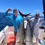 Two men on a fishing boat posing with several large tuna laid on the deck under a clear blue sky.