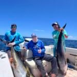 Three men on a boat posing with two large yellowfin tuna against a blue ocean and clear sky.