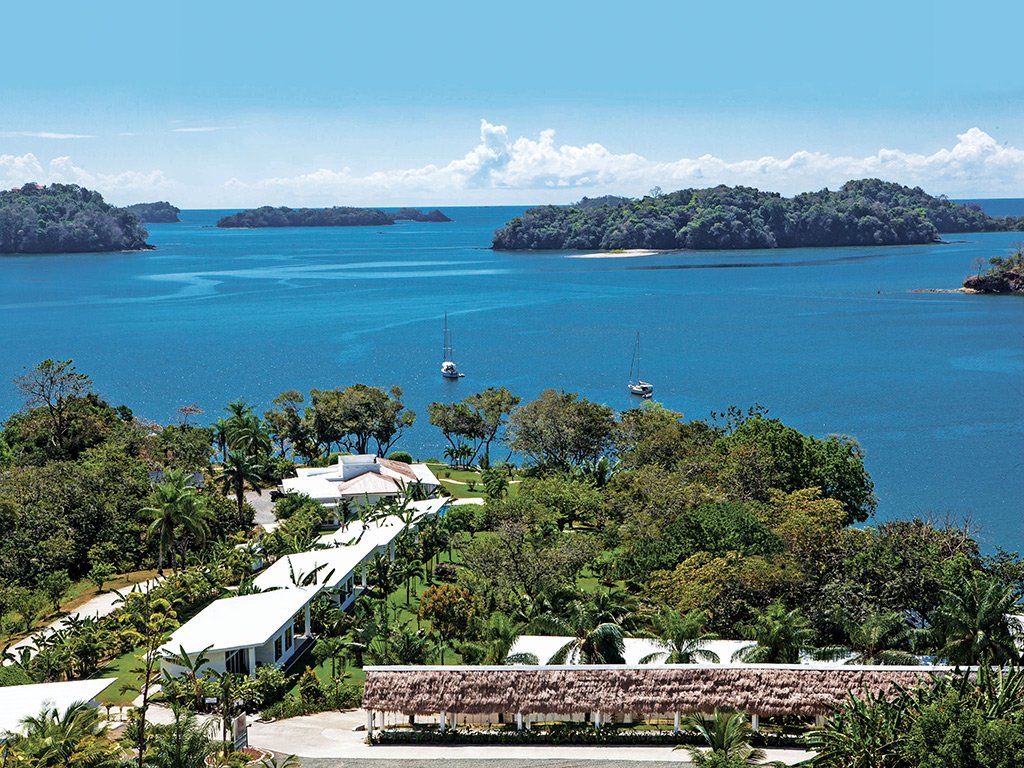 Aerial view of a tropical island resort with white buildings among palm trees along the shoreline, blue water, and two sailboats in the bay.