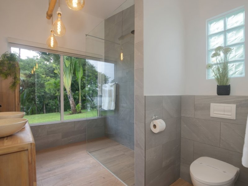 Bright modern bathroom with a glass-enclosed shower, wooden vanity with vessel sinks, and a view of a green garden through a large window.