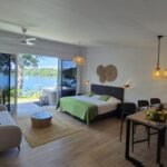 bdm18 - Panama Sport Fishing Charters Hotel room with a king-size bed in a green-spread, a beige sofa, and a wooden dining table; balcony doors open to a lake view with trees outside.