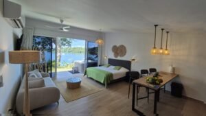 - Panama Sport Fishing Charters Hotel room with a king-size bed in a green-spread, a beige sofa, and a wooden dining table; balcony doors open to a lake view with trees outside.
