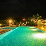 bdm2 - Panama Sport Fishing Charters Nighttime view of a lit pool with turquoise tiles, surrounded by lounge chairs, umbrellas, and a thatched-roof cabana area in a tropical resort.