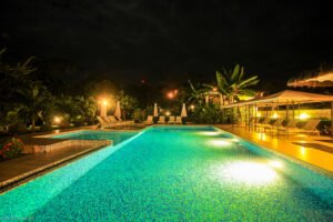 - Panama Sport Fishing Charters Nighttime view of a lit pool with turquoise tiles, surrounded by lounge chairs, umbrellas, and a thatched-roof cabana area in a tropical resort.
