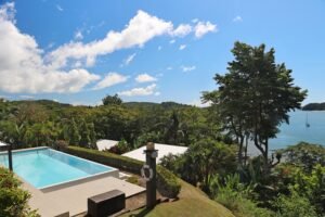 - Panama Sport Fishing Charters Swimming pool in a villa garden with lush tropical plants and a blue bay in the background, a small sailboat on the water.