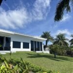 bdm24 - Panama Sport Fishing Charters White, single-story house with a covered patio on a green lawn, surrounded by palm trees under a bright blue sky.
