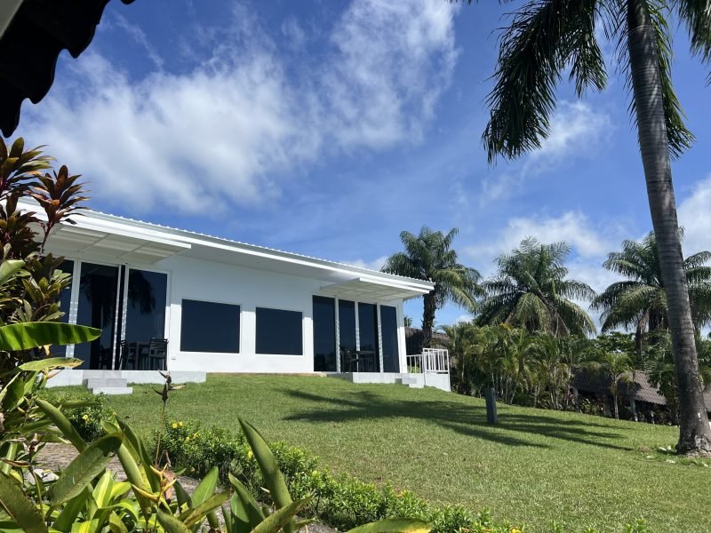 White, single-story house with a covered patio on a green lawn, surrounded by palm trees under a bright blue sky.