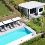 default - Panama Sport Fishing Charters Aerial view of a rectangular blue pool with white edge, surrounded by four cabanas with lounge chairs and small tables, on a sunny resort lawn.