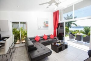 Bright modern living room with a black sectional sofa and red cushions, facing a coffee table, opening to a sunny patio by the water.