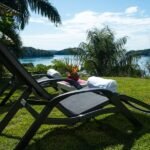 bdm28 - Panama Sport Fishing Charters Two dark lounge chairs on a grassy lawn facing a lake with palm trees and a blue sky, towels and a plant on a small table nearby.
