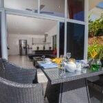 bdm29 - Panama Sport Fishing Charters Outdoor wicker dining table on a patio set for breakfast, with teapot, cups, juice, and a vase of green plants; glass doors open to a modern indoor kitchen and living area.