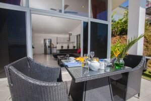 - Panama Sport Fishing Charters Outdoor wicker dining table on a patio set for breakfast, with teapot, cups, juice, and a vase of green plants; glass doors open to a modern indoor kitchen and living area.