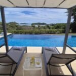 bdm30 - Panama Sport Fishing Charters Loungers under a cabana by a blue pool, with towels, a small table holding a bottle and glasses, and a distant ocean view on a sunny day.