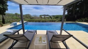 Loungers under a cabana by a blue pool, with towels, a small table holding a bottle and glasses, and a distant ocean view on a sunny day.