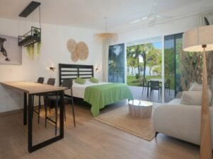 Bright, open-plan studio with a bed in green bedding, a seating area, and a balcony view of palm trees outside.