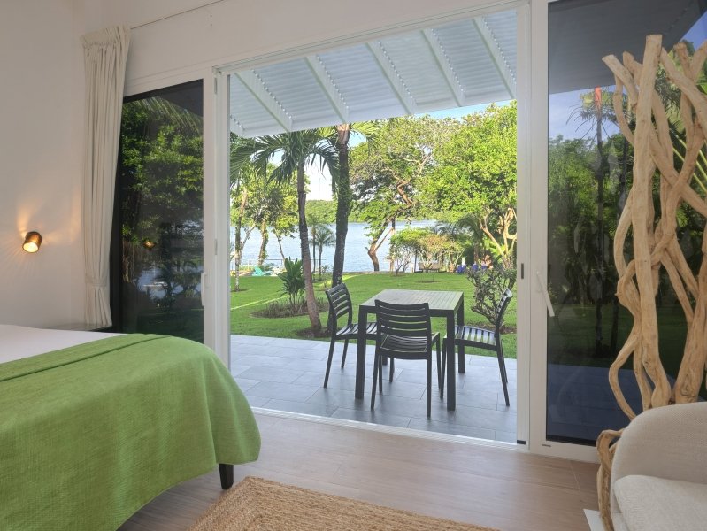 Bedroom with a green bedspread opens to a patio with a table and chairs, overlooking a lawn and lake.
