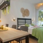 bmd12 - Panama Sport Fishing Charters Hotel suite with a breakfast-style table in the foreground, bed with green throw, and balcony view of palm trees and water.Recently designed interior with wicker wall art and plants above the table to the left.