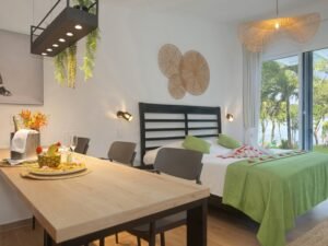 - Panama Sport Fishing Charters Hotel suite with a breakfast-style table in the foreground, bed with green throw, and balcony view of palm trees and water.Recently designed interior with wicker wall art and plants above the table to the left.