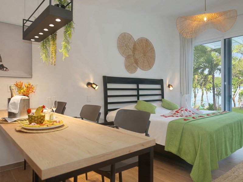 Hotel suite with a breakfast-style table in the foreground, bed with green throw, and balcony view of palm trees and water.Recently designed interior with wicker wall art and plants above the table to the left.