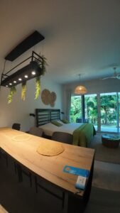 - Panama Sport Fishing Charters Long wooden dining table in a bedroom, with woven placemats and a black overhead light fixture with trailing greenery; bed with green bedding in the background.