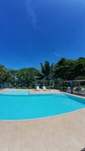 Outdoor pool deck with turquoise water, beige tiles, lounge chairs, and palm trees under a clear blue sky.