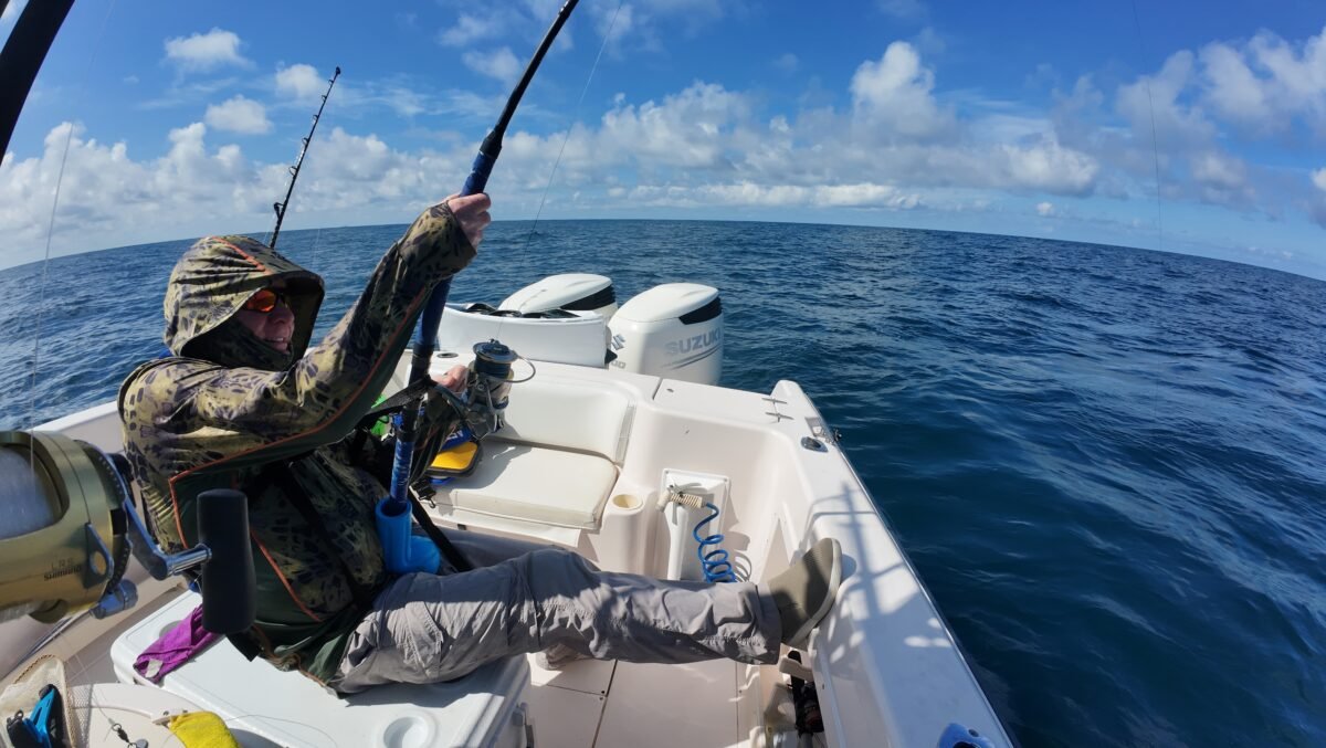 Fisherman in a camo hooded jacket fishing from a small white boat on a blue ocean under a bright sky.