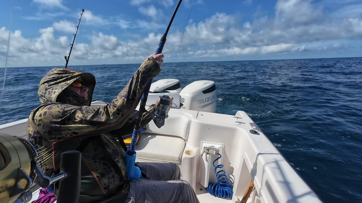 Fisherman in a camo jacket on a white boat, reeling in a fishing rod over the blue ocean under a partly cloudy sky.