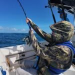 Fisherman on a boat wearing camouflage clothing, reeling in a bending fishing rod against a clear blue ocean and sky.