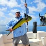 Man on a boat hoists a large yellow-green mahi-mahi with a gaff rod against a blue ocean and sky.