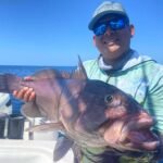 Man on a boat wearing a cap and sunglasses, proudly holding a large brown fish with the ocean behind him.
