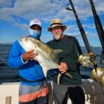 Two men on a boat pose with a large silver fish they just caught, with fishing rods and blue sky in the background.