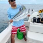 Man in a light blue shirt, red striped shorts, and a cap on a boat holding a large silver fish with tall dorsal spines, against the ocean backdrop.