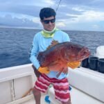 Man in a blue long-sleeve shirt and sunglasses holds a large red fish on a boat, orange gloves, open ocean behind him.