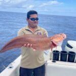 Man on a boat holding a large reddish-brown fish with its mouth open, ocean and blue sky in the background.