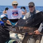 Three anglers on a boat posing with a large marlin; Panama Sport Fishing Charters banner visible, blue ocean behind.