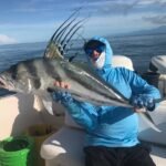 Person in a blue hoodie on a boat proudly holds a large silver fish with tall, ribbon-like dorsal fins against a calm sea backdrop