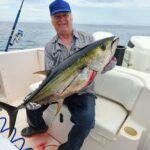 Man in a blue cap on a boat holding a large yellowfin tuna, with a fishing rod nearby and blood on the deck.