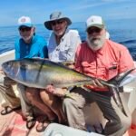 Three men on a boat proudly pose with a large yellowfin tuna against a calm ocean backdrop.