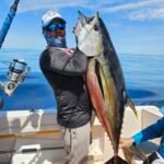 Man on a boat proudly holds up a large tuna caught while sporting a cap and sunglasses, calm blue ocean in the background.