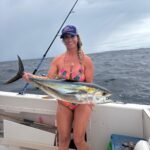 Smiling woman in a colorful bikini on a boat holding a large yellowfin tuna she just caught, fishing rod in the background at sea.