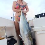 Smiling woman in a colorful bikini on a boat, holding a large yellowfin tuna by its tail in front of her.