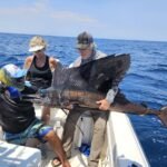 Three people on a fishing boat hold up a large sailfish, its long dorsal fin spread, against a blue ocean backdrop.