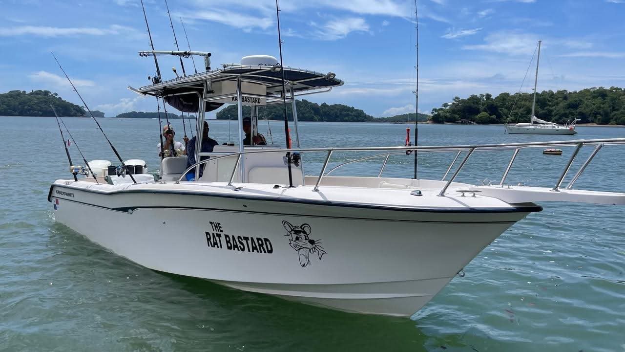 White fishing boat named 'The Rat Bastard' with a cartoon rat logo on calm blue water.